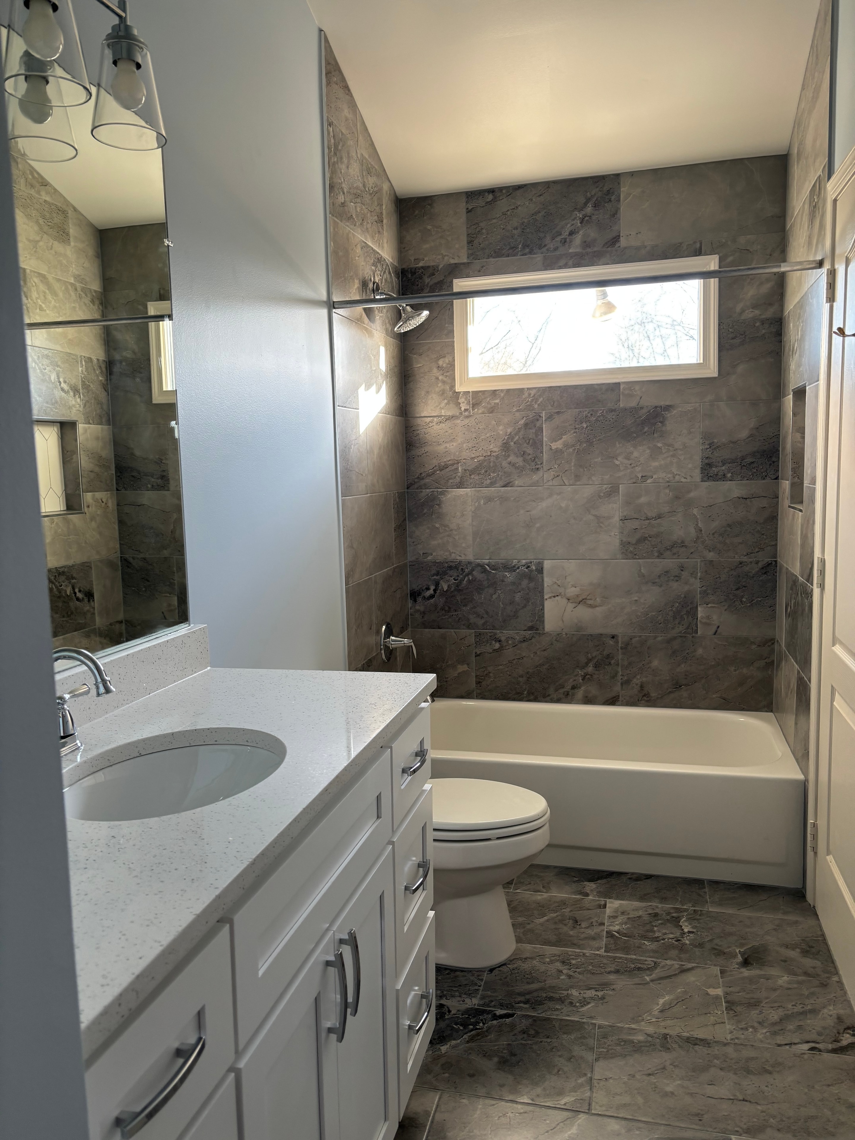 Bright bathroom with gray walls, white tile shower, window, and gray floor, lit by natural light