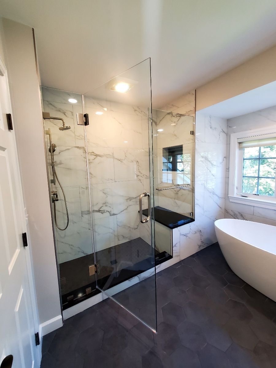 Modern bathroom with glass shower, marble walls, dark floor, and freestanding tub near a window